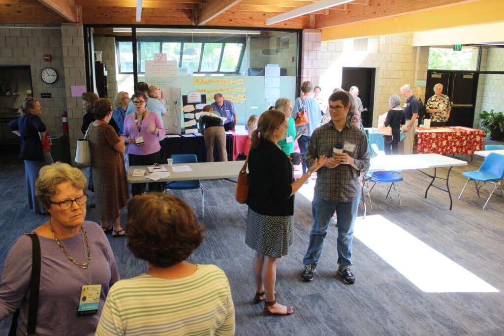 This is a photo of Presidents Hall at First UU of Richmond with several people standing in small groups, engaged in conversation and holding cups of coffee. Some people are standing at an information table near the back of the picture.