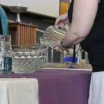 This is a picture of the unseen people preparing the water communion at a purple table; somebody is pouring a picture of water into a clear bowl of water; the chalice is in the background.