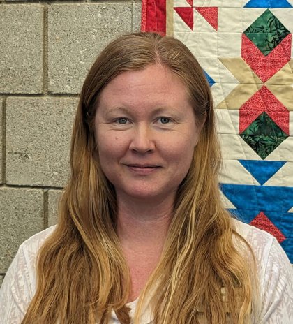 LeighAnn Luscan - Dir Lifespan Faith Formation, A person with long hair in front of a colorful quilt and brick wall.