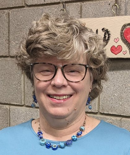 Person with curly gray hair and glasses, wearing a blue top and jewelry, against a brick wall background.