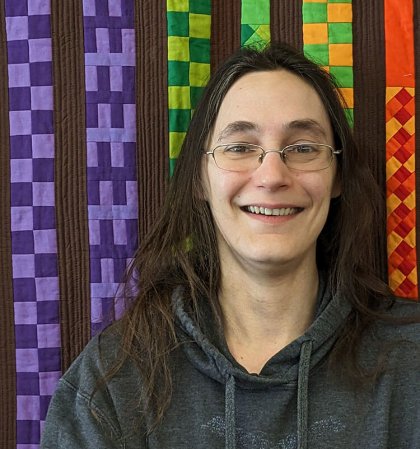 Person wearing glasses and a gray hoodie, with a colorful quilted background.