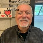 Frank Corsoro, Board Member, Caucasian male with gray follicles frolicking; a U-shaped grey mustache and beard; wearing a brown button down shirt with a black t-shirt underneath