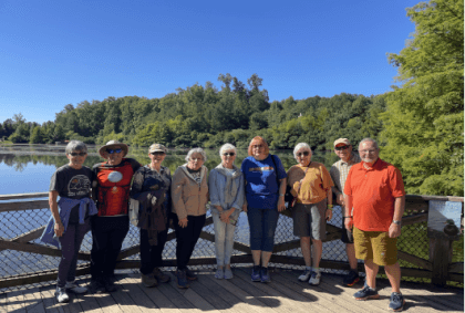 9 neighbors on a wooden dock on the Mines Walk; river and green trees in the background