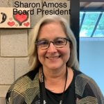 Sharon Amoss Board President. Caucausian woman with gray hair; wearing dark-rimmed glasses and a dark gray sweater with different dark hues; landyard around her neck 