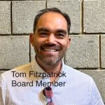 Tom Fitzpatrick Board Member, a Caucasian man with black hair, mustache and beard wearing a white polo shirt with clasped to his shirt