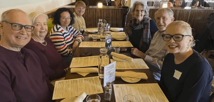 This is a picture of eight church members from first UU having lunch at a table. There are napkins and glasses are on the table. Four people are seated on the left side. Three people are seated on the right side.