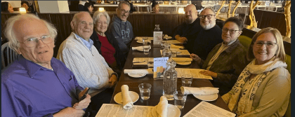 This is a picture of eight church members from first UU having lunch at a table. There are napkins and glasses are on the table. Four people are seated on each side.