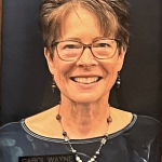 Portrait of a person smiling with glasses and short hair, wearing a dark top and necklace.