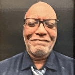 Person wearing glasses and a tie against a dark background.