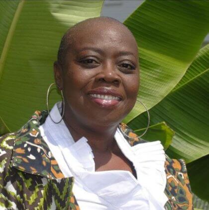 BIPOC Person standing in front of large banana leaves, wearing a patterned jacket and white top.