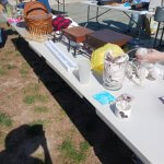 Outdoor table at a yard sale with assorted items like baskets, light tubes, and glassware, under bright sunlight.