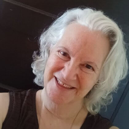Late middle ages white woman with white wavy hair that is cut above her shoulders, smiling at the camera in a brown tank top