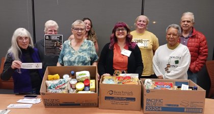 Group of mixed aged people standing behind a wood table with three boxes on it that are full of dry good food stuffs. 